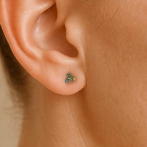 May include: A close-up shot of a woman's ear, showcasing a small stud earring. The earring is adorned with three teal gemstones, set in a triangular formation. The skin is fair, and the hair is styled up and away from the ear.