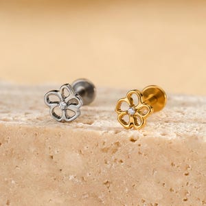 May include: Two flower-shaped stud earrings. One is silver-toned, the other is gold-toned with a small, clear gem in the center. The earrings are displayed on a textured, light brown surface, with a soft, blurred background.
