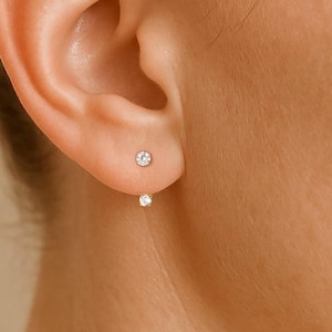 May include: Close-up of a person's ear with two small, round, sparkling stud earrings. The top earring is a single, small, clear gemstone. The second earring is a similar gemstone, positioned below the earlobe. The skin tone is fair.