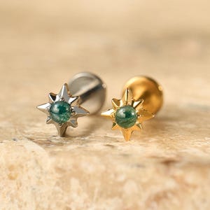 May include: Two stud earrings, one silver and one gold. Each has a starburst design with a small, round, green gemstone in the centre. The earrings are displayed on a textured, light brown surface.