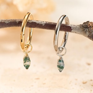 May include: A pair of hoop earrings, one gold and one silver, each with a small, green and white stone pendant. The earrings are displayed on a small branch against a neutral background, showcasing their design and color contrast.