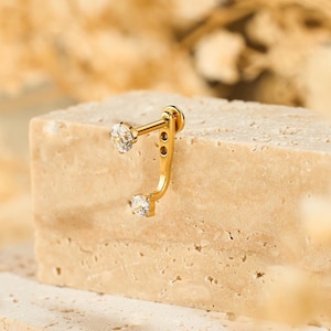 May include: A gold-colored ear cuff with two clear, round gemstones. The earring is displayed on a light beige stone block, with dried flowers in the background. The earring has a curved design.