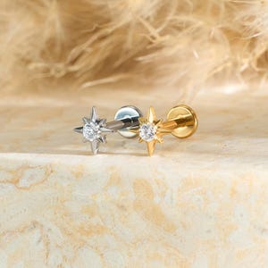 May include: Two star-shaped earrings, one silver and one gold, each with a central clear gemstone. The earrings are displayed on a light-colored, textured surface, with a blurred, feathery background.