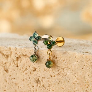 May include: A gold-colored earring with a cluster of small, green gemstones and a dangling, round green gem. The earring is set against a neutral, textured background, highlighting its delicate design and color.