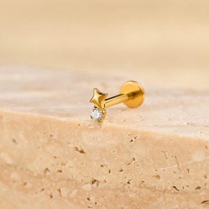 Könnte beinhalten: Goldfarbener Ohrring in Sternform mit kleinem, klarem Edelstein. Der Ohrring wird auf einer strukturierten, hellbraunen Oberfläche präsentiert. Der Ohrring ist für ein Lippen- oder Ohrpiercing konzipiert.