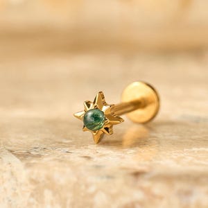 May include: A gold stud earring with a starburst design. The starburst has a small, round, green gemstone at its centre. The earring is set against a neutral, textured background.