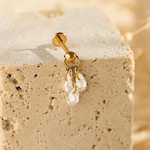 May include: A gold-coloured earring featuring a small, flat top and a cluster of three clear, teardrop-shaped crystals. The earring is displayed on a textured, beige stone block, with a soft, blurred background.