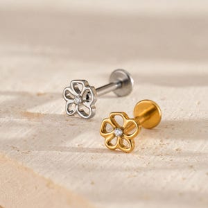 May include: Two flower-shaped labret earrings. One is silver-colored with a clear gem in the center, and the other is gold-colored with a clear gem in the center. The earrings are displayed on a light-colored, textured surface.