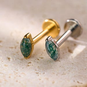 May include: Two stud earrings with marquise-shaped green gemstones. One earring has a gold-colored post and setting, while the other has a silver-colored post and setting. The earrings are displayed on a textured, light-colored surface.