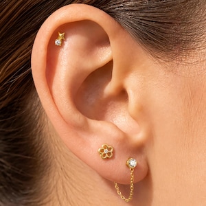 May include: A gold ear piercing set featuring a small star stud with a clear stone, a flower-shaped stud, and a chain-linked stud with a clear stone. The jewellery is worn in the upper cartilage and the earlobe, showcasing a polished gold finish.