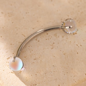 May include: Curved eyebrow piercing jewelry with a silver-colored, curved barbell. Each end features a clear, iridescent, round gem. The jewelry is displayed on a light beige, textured surface.