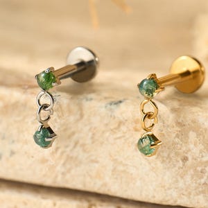 May include: Two ear piercing studs. One stud is silver-colored, the other is gold-colored. Each stud features a small, round, green gemstone at the top and a dangling green gemstone. The earrings are displayed on a light-colored, textured surface.