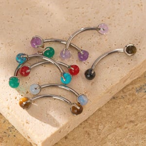 May include: A collection of curved eyebrow rings with various coloured gemstone beads. The jewellery features purple, green, red, turquoise, and brown beads. The metal is silver-toned, and the gemstones are round.