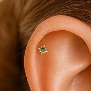 May include: A close-up of an ear with two small gold earrings. Each earring has a teal gemstone in a starburst setting. The earrings are placed on the upper ear. The earrings are a delicate accessory.