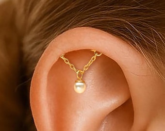 Könnte beinhalten: Ein goldfarbener Ohrschmuck mit einer feinen Kette und einem kleinen, runden, perlenartigen Anhänger. Der Schmuck befindet sich am oberen Ohrknorpel und wirkt zart. Der Hintergrund ist unscharf, wodurch der Fokus auf dem Ohrring liegt.