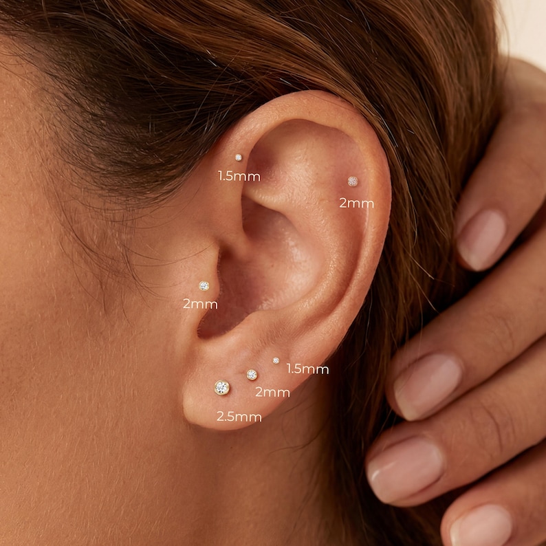 May include: An ear adorned with several small stud earrings in gold. The earrings are of varying sizes, including 1.5mm, 2mm, and 2.5mm. The image shows the ear with the earring sizes clearly labelled in millimeters.