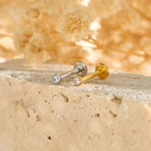 May include: Two small stud earrings, one silver and one gold. Each has a clear, round gemstone. The earrings are displayed on a textured, light-coloured surface, with a blurred floral background.