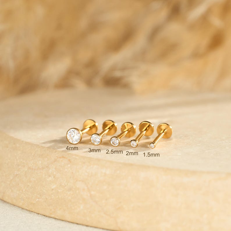 May include: A selection of gold stud earrings, each featuring a clear, round gemstone. The earrings are of varying sizes, with the largest measuring 4mm and the smallest 1.5mm. The earrings are displayed on a light-coloured surface.