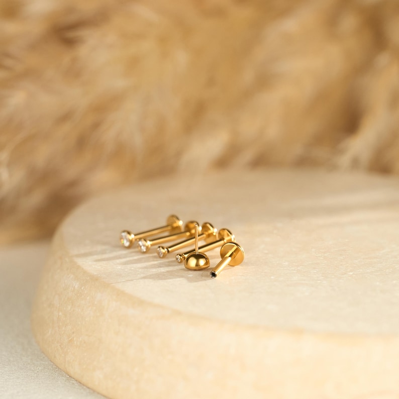May include: A selection of gold-coloured stud earrings with different designs. Some studs have small, clear gemstones, while others have a smooth, rounded appearance. The earrings are arranged on a light-coloured, round surface.