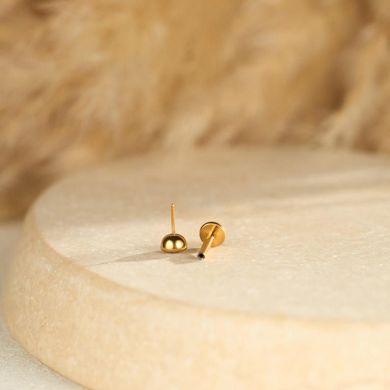 May include: Two gold earrings are shown on a light-coloured, round surface. One earring has a small, round stud, and the other has a flat, circular stud. The background is a soft, blurred tan colour.