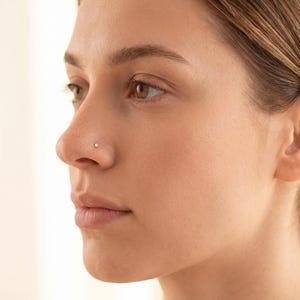 May include: A profile view of a person's face, highlighting the nose. A small, round, silver nose stud is set in the nostril. The skin tone is fair, and the background is a soft, neutral colour.