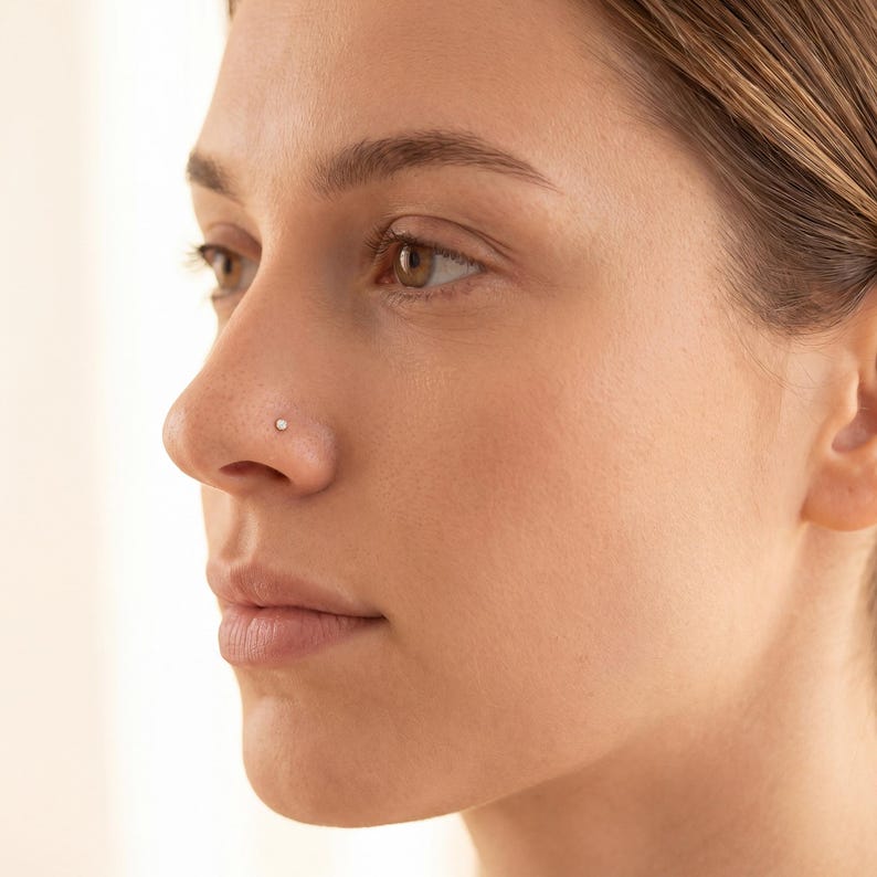 May include: A profile view of a person's face, highlighting the nose. A small, round, silver nose stud is set in the nostril. The skin tone is fair, and the background is a soft, neutral colour.