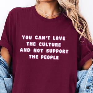 Puede incluir: Una camiseta color granate con el texto "You can't love the culture and not support the people" en letras blancas con contorno rosa.