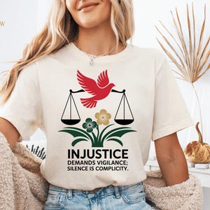 Op de afbeelding: Crèmekleurig T-shirt met een afbeelding van een rode duif met een weegschaal, het woord "INJUSTICE" en de tekst "DEMANDS VIGILANCE; SILENCE IS COMPLICITY." Het ontwerp bevat groene en gouden bloemelementen.