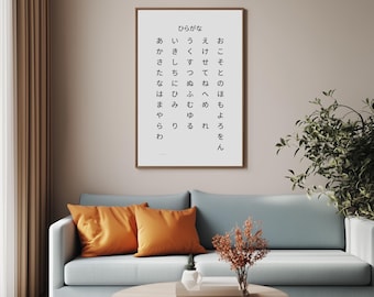 May include: A white framed print with black text on a white background. The text is in Japanese and reads "hiragana" and then lists the Japanese alphabet in hiragana. The print is hanging on a light beige wall above a light blue sofa with two orange throw pillows.