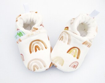 May include: A pair of white baby booties with a brown, yellow, and orange rainbow pattern. The booties have a soft, plush lining and a pull-on design.