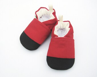 Può includere: Un paio di scarpine rosse per bambini con suole nere. Le scarpe hanno un interno morbido e bianco e un piccolo anello sul retro. Le scarpe sono realizzate in un materiale simile alla tela.