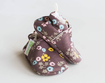 May include: A pair of brown baby booties with a floral print featuring pink, yellow, and blue flowers. The booties are made of soft fabric and have a comfortable fit.