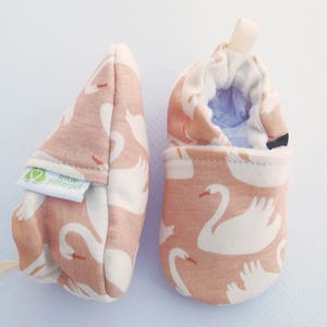 May include: A pair of pink baby shoes with a white swan pattern. The shoes have a soft, comfortable fabric and a secure closure. The shoes are made by the brand "Little Pitter Pat"