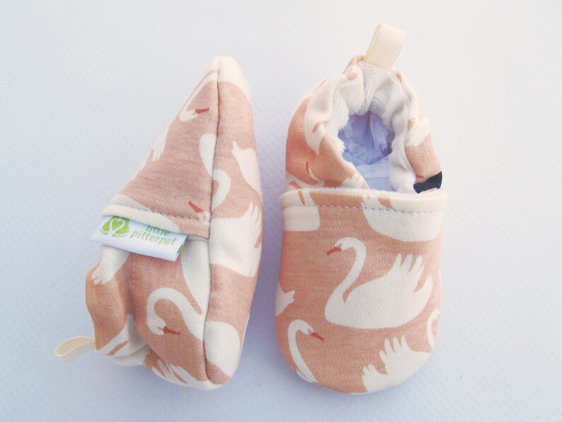 May include: A pair of pink baby shoes with a white swan pattern. The shoes have a soft, comfortable fabric and a secure closure. The shoes are made by the brand "Little Pitter Pat"