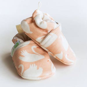 May include: A pair of pink baby shoes with a white swan pattern. The shoes are made of soft fabric and have a comfortable fit.