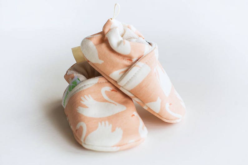 May include: A pair of pink baby shoes with a white swan pattern. The shoes are made of soft fabric and have a comfortable fit.