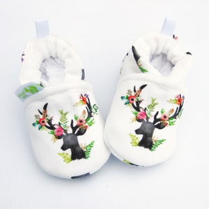 May include: A pair of white baby booties with a black deer head design adorned with colorful floral accents. The booties have a soft, plush interior and a small loop at the back for easy wear. The design includes green leaves, pink, orange, and yellow flowers.