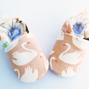 May include: A pair of baby booties with a swan pattern. The booties are made of a soft, peach-colored fabric with white swans printed on them. The booties have a light blue lining and a white elastic band at the top.