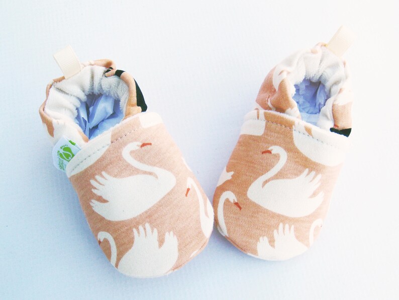 May include: A pair of baby booties with a swan pattern. The booties are made of a soft, peach-colored fabric with white swans printed on them. The booties have a light blue lining and a white elastic band at the top.