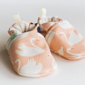 May include: A pair of pink baby booties with a white swan pattern. The booties have a soft, padded lining and a flexible sole.