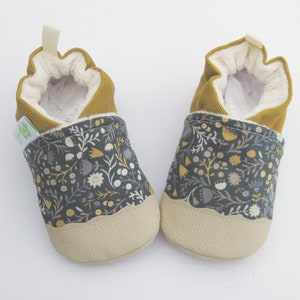 Pu&ograve; includere: Un paio di scarpine per neonati con un motivo floreale in tonalit&agrave; di grigio, giallo e bianco. Le scarpine hanno un polsino giallo senape e una suola beige.