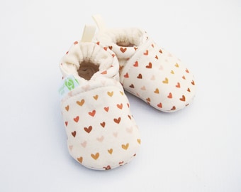 May include: A pair of cream-colored baby booties with a brown, pink, and orange heart pattern. The booties have a soft, fuzzy lining and a fold-over cuff.