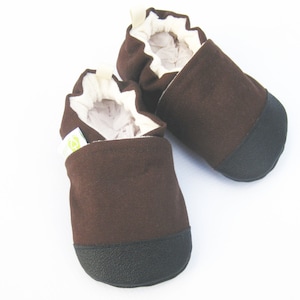 May include: A pair of brown baby shoes with black soles and off-white lining. The shoes are made of a soft fabric and have a simple, classic design. The shoes are designed for infants and toddlers.