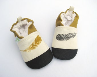 May include: A pair of baby booties with a black and white feather design. One bootie has a gold feather and the other has a black feather. The booties are made of a soft, canvas material and have a black rubber sole.