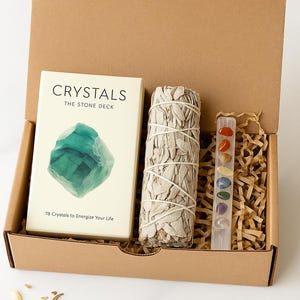 Pode incluir: Uma caixa de cartão castanha contendo um baralho de cristais, um maço de sálvia e um conjunto de cristais coloridos. O baralho de cristais está rotulado como "CRYSTALS THE STONE DECK" com o texto "78 Crystals to Energize Your Life". O maço de sálvia é amarrado com barbante branco.