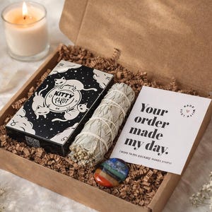 May include: A brown gift box with a Kitty Tarot deck, sage bundle, chakra stone, and a lit candle. The tarot box is black with white cat designs. A card reads "Your order made my day."