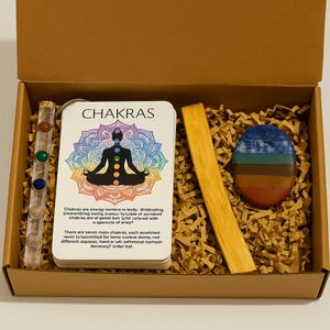 Op de afbeelding: Een doos met een heldere kristallen toverstaf met gekleurde stenen, een kaartspel met het woord "CHAKRAS" en een chakra-diagram, een houten stok en een kleurrijke ovale steen. De doos is gevuld met bruin papier.