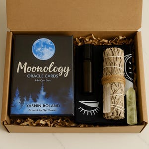 Puede incluir: Una caja de regalo que contiene una baraja de "Moonology Oracle Cards", una pequeña botella de aceite esencial, un manojo de salvia, un cristal y una tela negra con un diseño de ojo. La caja de cartas es negra con un gráfico de luna azul y el texto "Moonology Oracle Cards".
