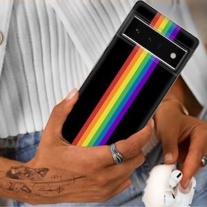 Puede incluir: Una funda de teléfono negra con un diseño de rayas arcoíris. La funda la está sujetando una persona que lleva un suéter blanco y pantalones vaqueros azules.