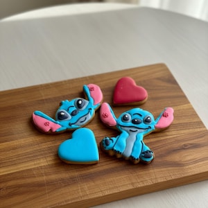 Pode incluir: Uma tábua de corte de madeira exibe biscoitos de açúcar decorados. Estão presentes dois biscoitos azuis com personagens com orelhas cor-de-rosa e um biscoito em forma de coração. Um biscoito azul em forma de coração e um biscoito vermelho em forma de coração também são visíveis.